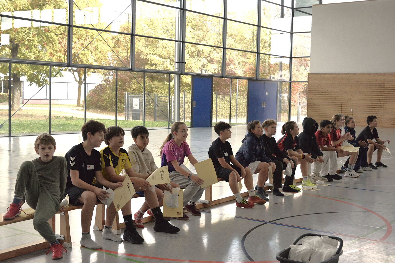 Schülerinnen und Schüler sitzen aufgereiht auf Bänken in der Sporthalle und warten, hinter ihnen die Glasfront.