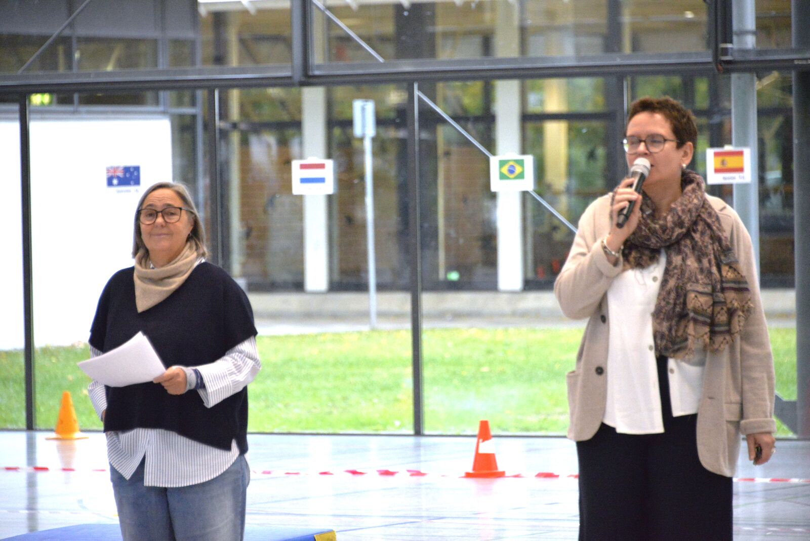 Zwei Frauen bei der Begrüßung in der Sporthalle: Eine spricht ins Mikrofon, die andere hält Notizzettel. Im Hintergrund hängen Länderflaggen an der Glasfront.