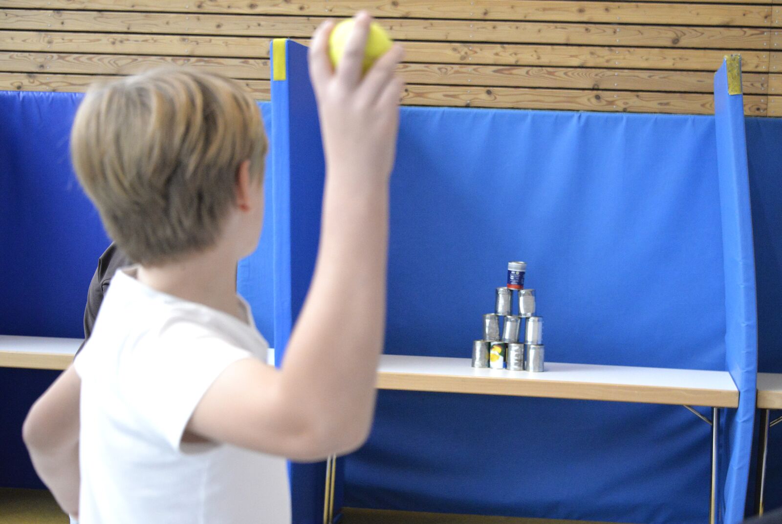 Ein Junge holt mit einem Ball zum Wurf aus und zielt auf eine Dosenpyramide vor blauen Matten.