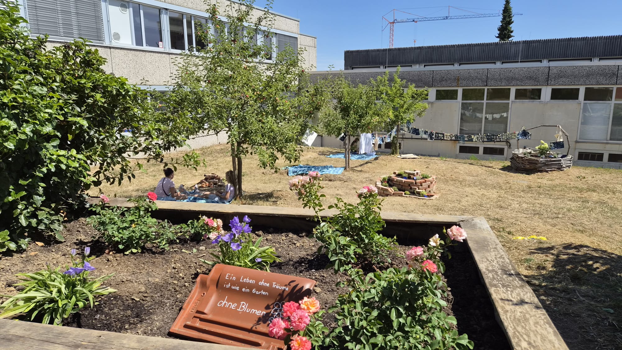 Hochbeet mit blühenden Rosen und Glockenblumen im Schulgarten. Vorne liegt ein Dachziegel mit der Aufschrift „Ein Leben ohne Träume ist wie ein Garten ohne Blumen“. Im Hintergrund sitzen zwei Schülerinnen auf einer Decke unter Obstbäumen, dazu Kunstobjekte aus Ziegeln und Holz und eine Leine mit blauen Cyanotypie-Bildern.